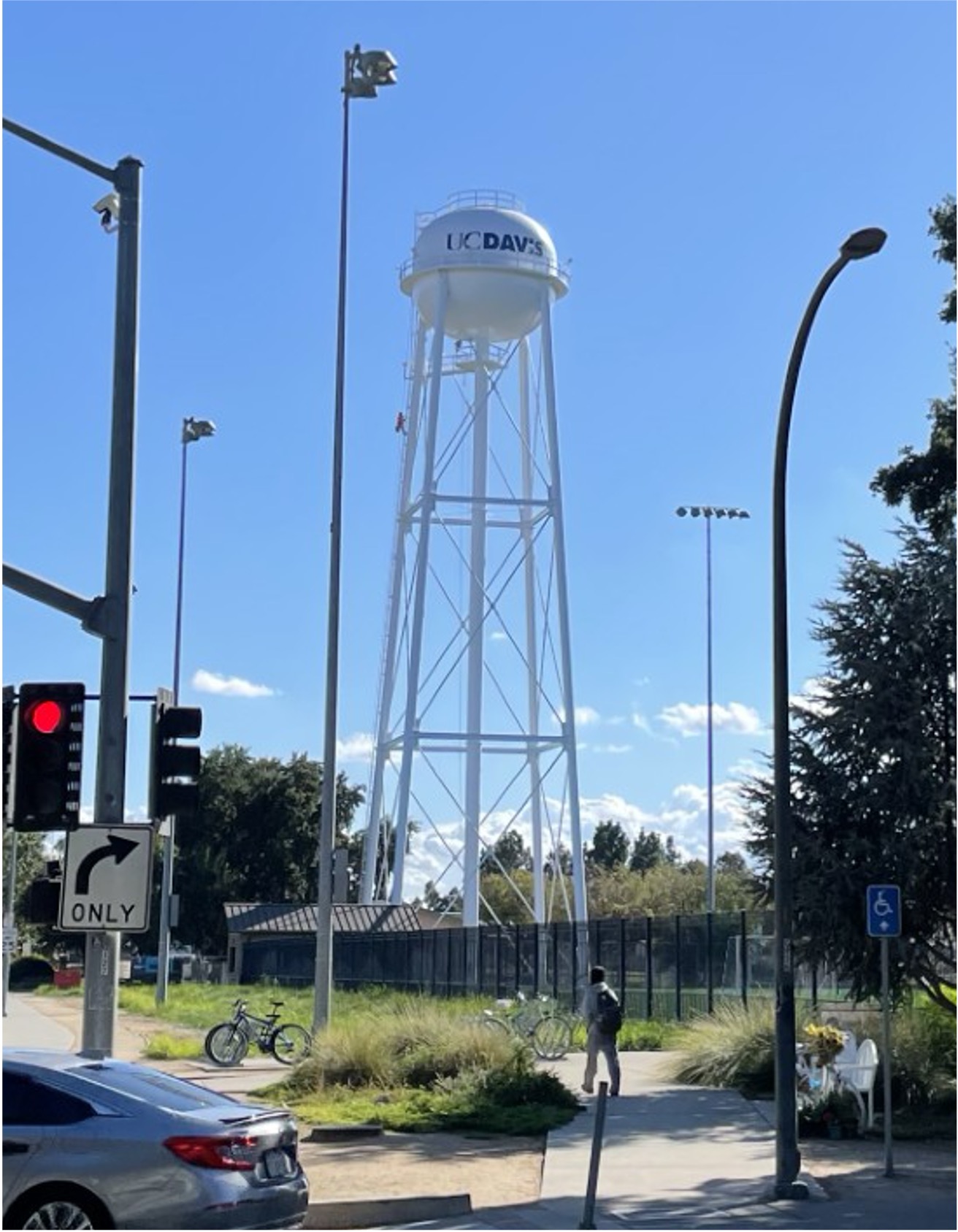 A tall, white steel water tower stands under a clear blue sky near a street intersection on the UC Davis campus. The tower features the blue “UC Davis” wordmark painted on the tank. Bicycles, a pedestrian, and traffic lights are visible in the foreground.