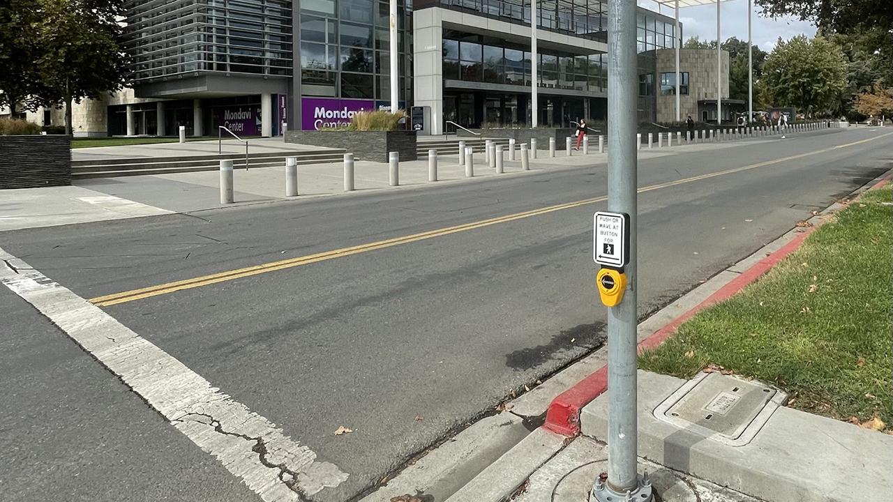 A new Accessible Pedestrian Signal (APS) device with a push button and touch-free wave sensor is mounted on a pole at a crosswalk in front of the UC Davis Mondavi Center.