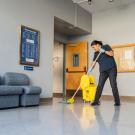 A UC Davis custodial services staff member wearing gloves mops a polished floor in a campus building lobby, with a yellow mop bucket nearby, seating along the wall, and bulletin boards and doors in the background.