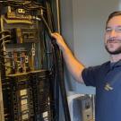 A smiling technician works on a circuit breaker panel, surrounded by wires and equipment.