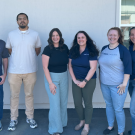 Group photo of seven colleagues standing by a white building, smiling.