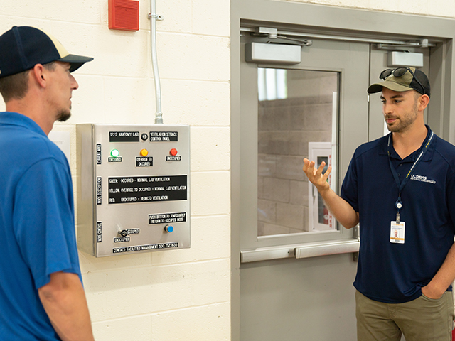 Image of Gary Harmon, a HVAC Controls Technician (left), and Sam Cole, one of our Energy Engineers (right), with the control panel they’ve created together. Gary was responsible for the electrical design and efficient wiring system. The green occupied light signifies active ventilation, exhaust and temperature control. According to Sam, the controls panel can help save the building $6000-7000/year in energy by ventilating when the room is in use instead of leaving the lab room ventilation on 24/7.